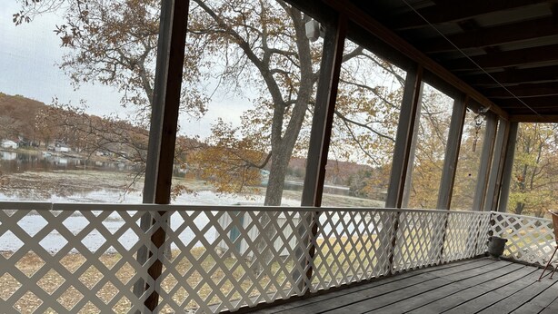Une terrasse en bois couverte, dotée d’une balustrade en treillis blanc, d’un tapis et offrant une vue sur un lac et des arbres.