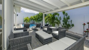 A covered outdoor seating area with wicker furniture, a pool, and tropical plants. - NEW 5BR Villa w/ PrivatePool 3min to Eagle Beach (Noord)