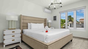 A hotel room with a large bed, a bedside table with a lamp, an air conditioning unit, and a window with a view of palm trees. - NEW 5BR Villa w/ PrivatePool 3min to Eagle Beach (Noord)