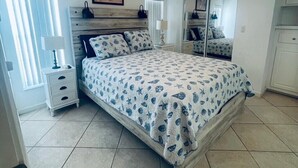 A bedroom with a bed, a nightstand, a lamp, a ceiling fan, a window with blinds, a door, and a mirror. - Cozy vacation home (Jensen Beach)