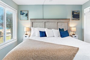 A bedroom with a bed, two bedside tables, two lamps, a window with blinds, and two wall-mounted paintings. - Ocean View Beachfront +In/Outdoor Pools + Sauna! (Gulf Shores)