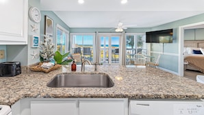 A kitchen with granite countertops, a stainless steel sink, and a dishwasher. There is a dining area with a table and chairs, and a view of the ocean through large windows.