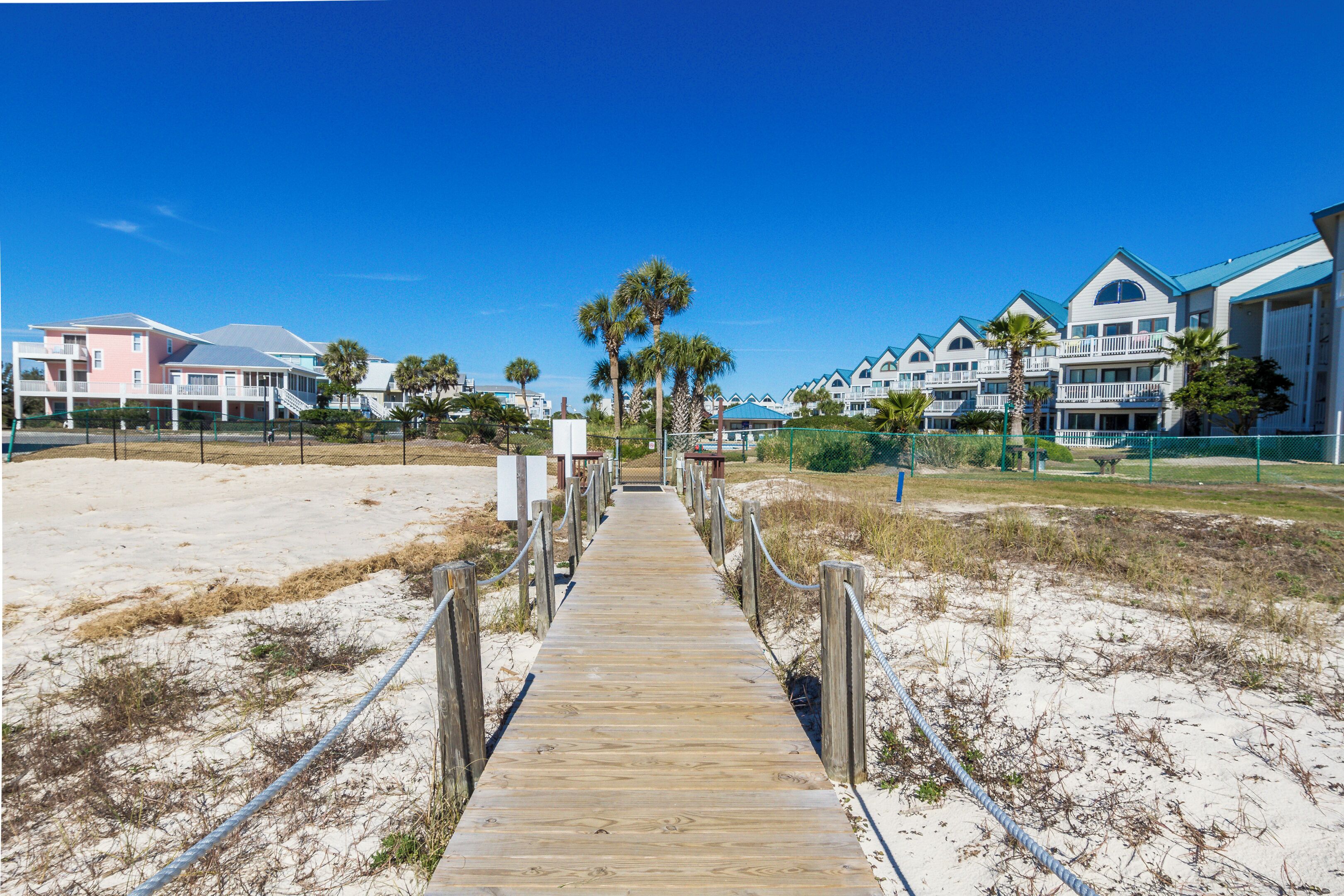 A wooden boardwalk leading to a beachfront hotel with multiple balconies and palm trees.