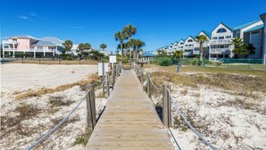A wooden boardwalk leading to a beachfront hotel with multiple balconies and palm trees.