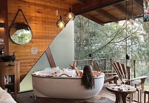 Interior de madera con una bañera llena de hielo y pétalos de rosa, una mesa de madera con una copa de vino y un ventanal con vistas a árboles.