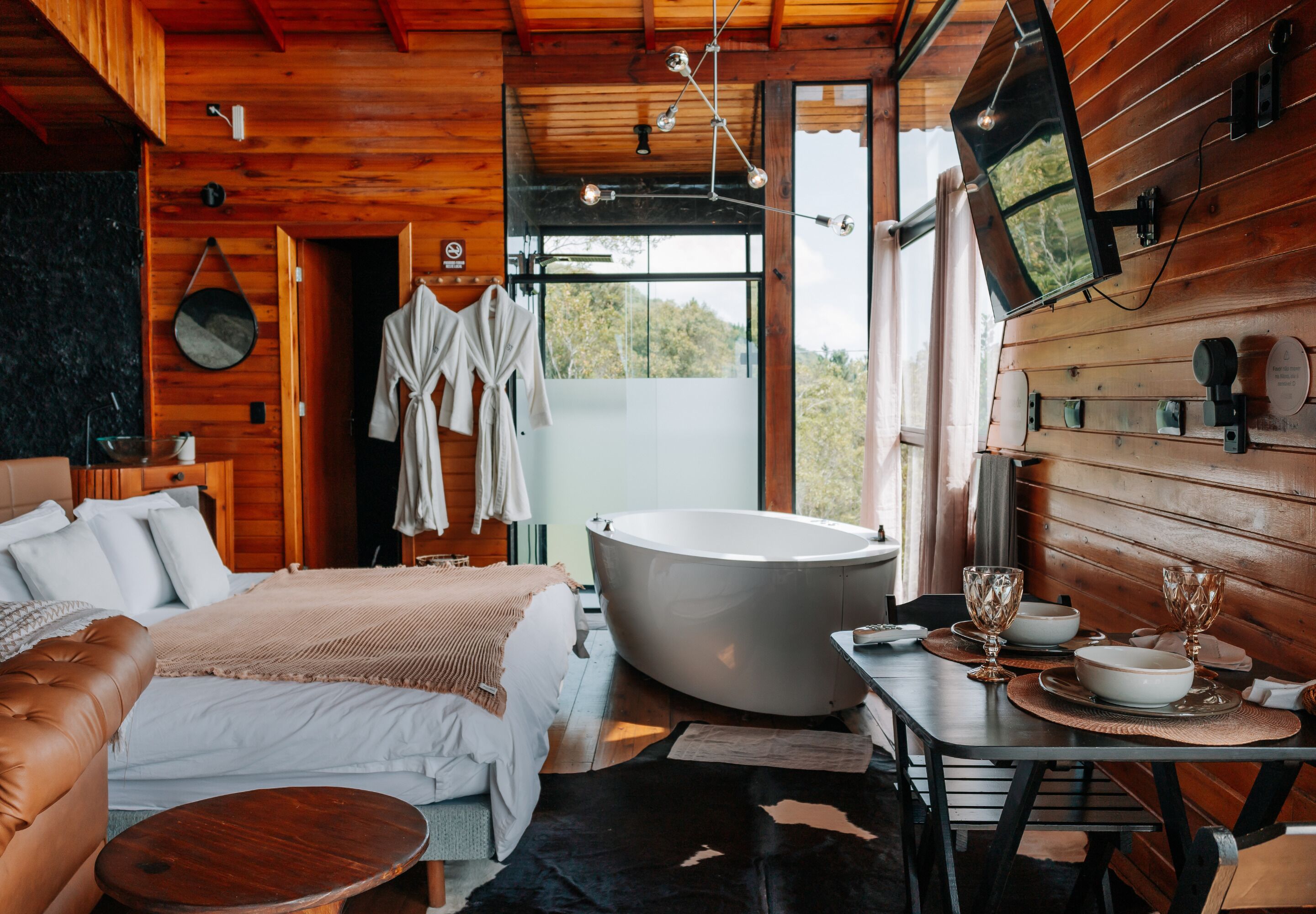 L'intérieur d'un chalet en bois comprend une grande baignoire, un lit avec une literie blanche, une table à manger avec un set de table et un téléviseur fixé au mur.