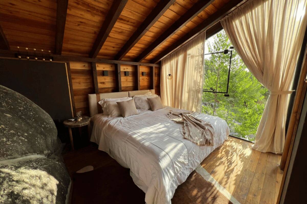 A wooden cabin bedroom with a bed, a small table, and a view of greenery through a window.