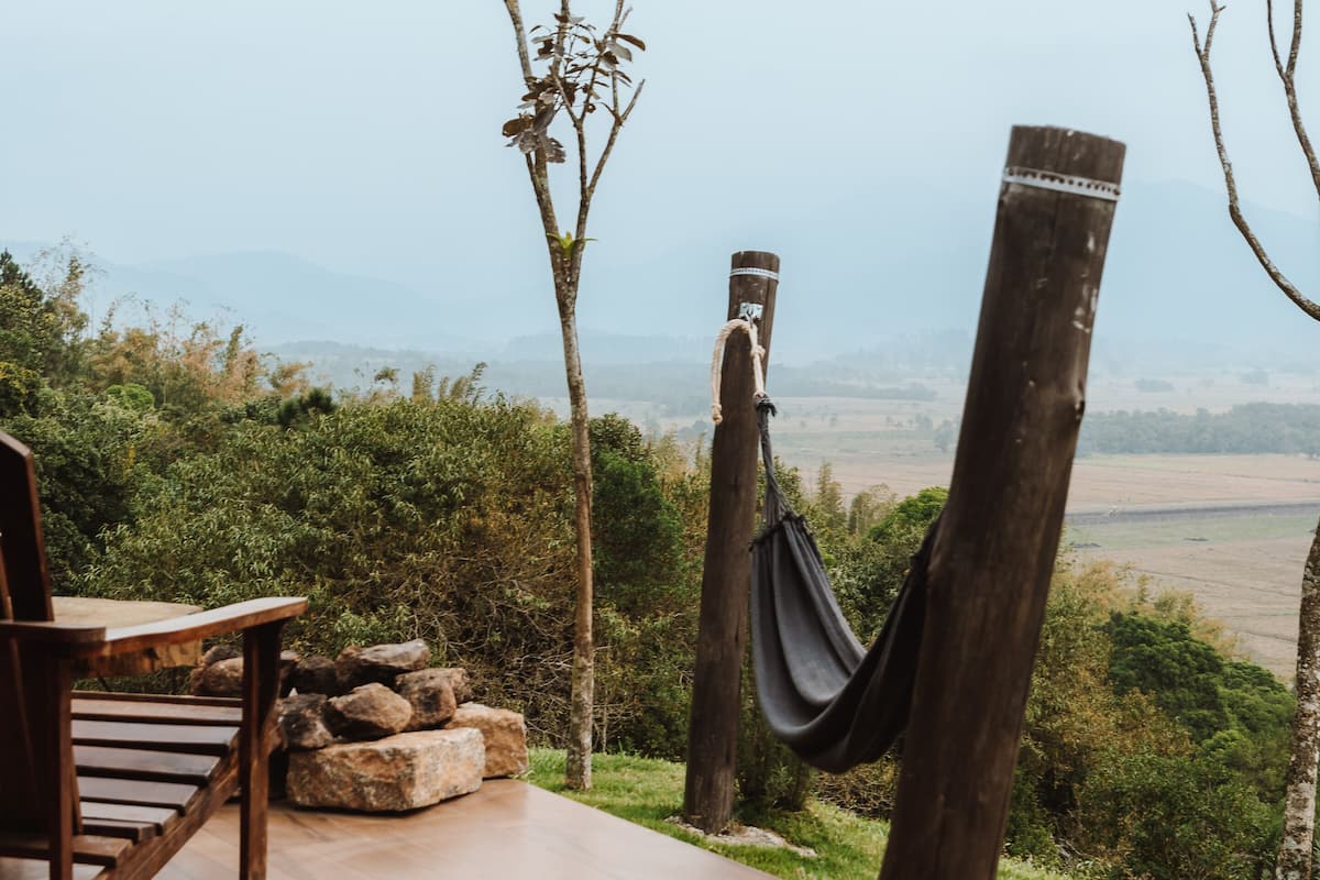 A wooden deck with a hammock, a chair, and a stone bench overlooking a valley.