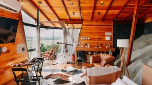 Interior de una cabaña de madera con comedor, sofá y vistas al exterior.