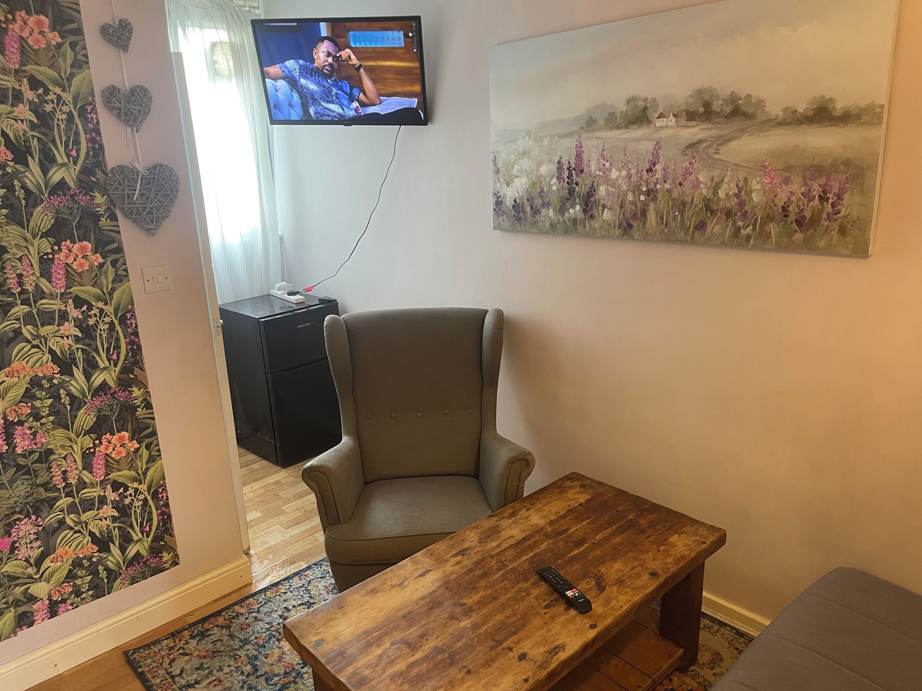Ett vardagsrum med en tv som är monterad på väggen, en beige fåtölj, ett soffbord i trä och en väggtapet med blommönster.
