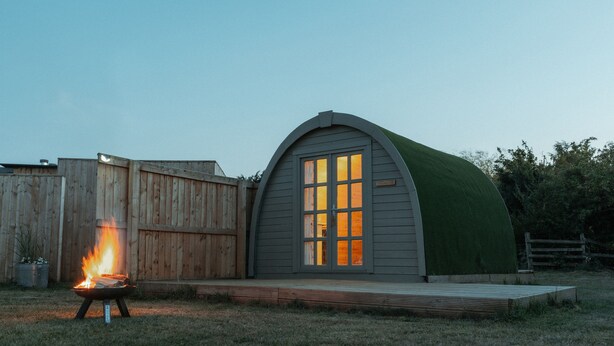 A small, curved wooden structure with a green roof, a fire pit in front, and a wooden deck. - Glamping in Wiltshire the Green Knoll is a charm (Chippenham)