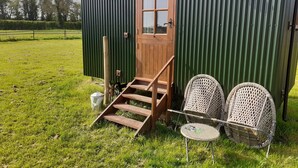 A green metal shed with a wooden door, a set of wooden steps leading up to it, and two wicker chairs outside. - Glamping in Wiltshire the Green Knoll is a charm (Chippenham)
