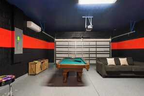 A pool table in a room with a garage door, a couch, and a cabinet with pool cues.