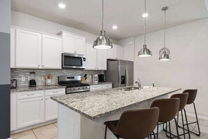 Cocina moderna con armarios blancos, electrodomésticos de acero inoxidable, encimeras de granito e iluminación colgante.