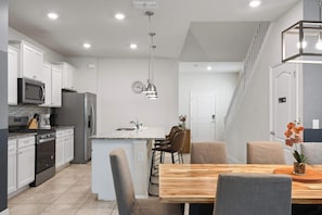 A modern kitchen with white cabinetry, a stainless steel refrigerator, a microwave, and a stove. There is a dining area with a wooden table and chairs, and a staircase leading to another level.