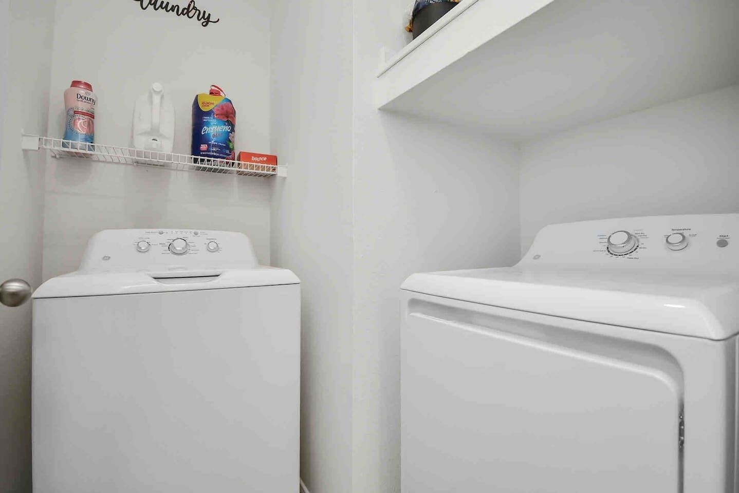 A laundry area with two white washing machines and a shelf with laundry detergent bottles.