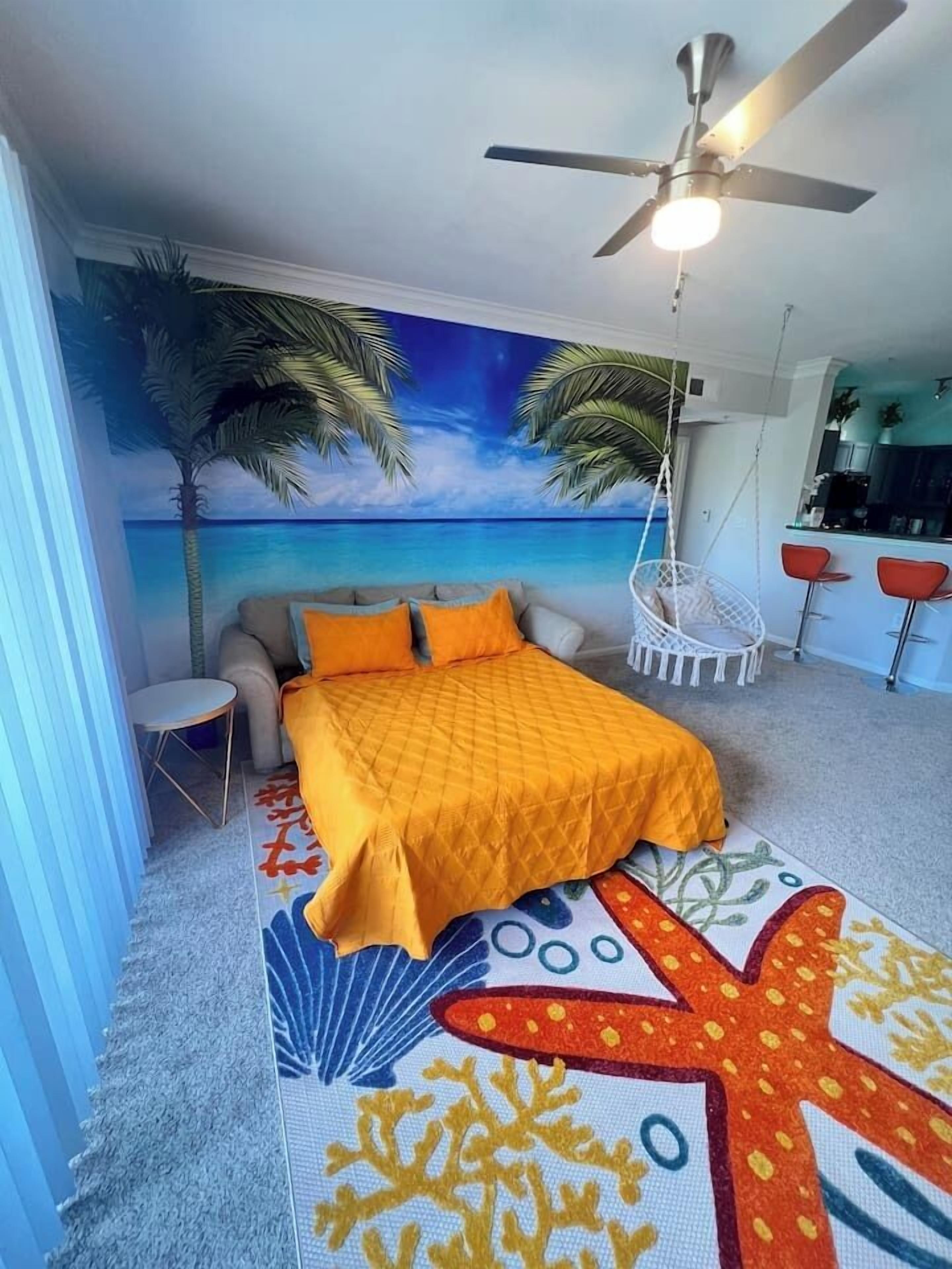 Camera da letto con un letto, una ventola a soffitto, un affresco raffigurante una spiaggia e un angolo cottura con sgabelli da bar.
