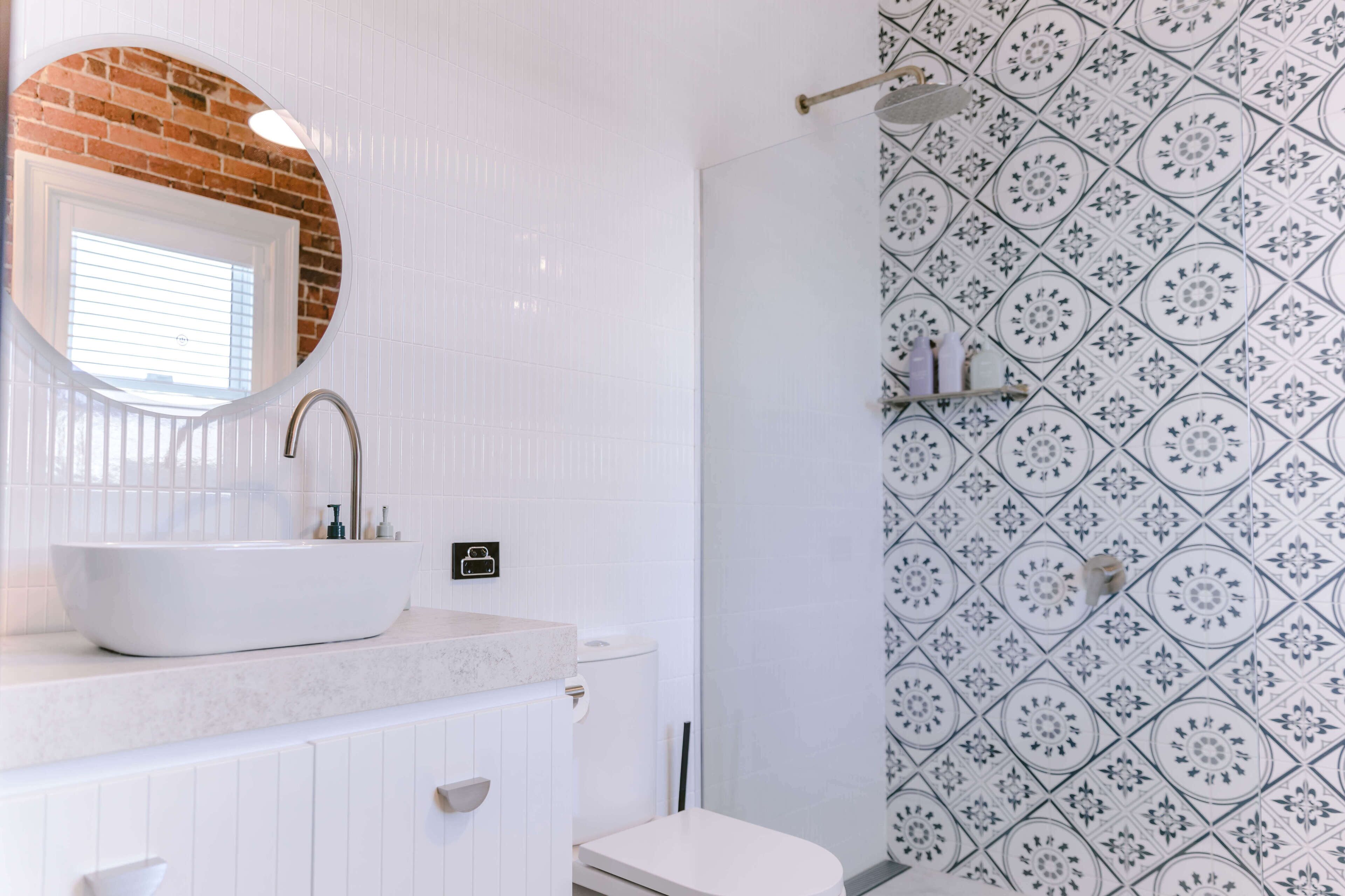 Un baño con un lavamanos blanco, un espejo redondo y un inodoro. Las paredes tienen un diseño de baldosas con motivos.