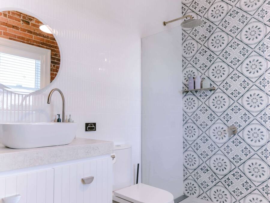 Un baño con un lavamanos blanco, un espejo redondo y un inodoro. Las paredes tienen un diseño de baldosas con motivos.
