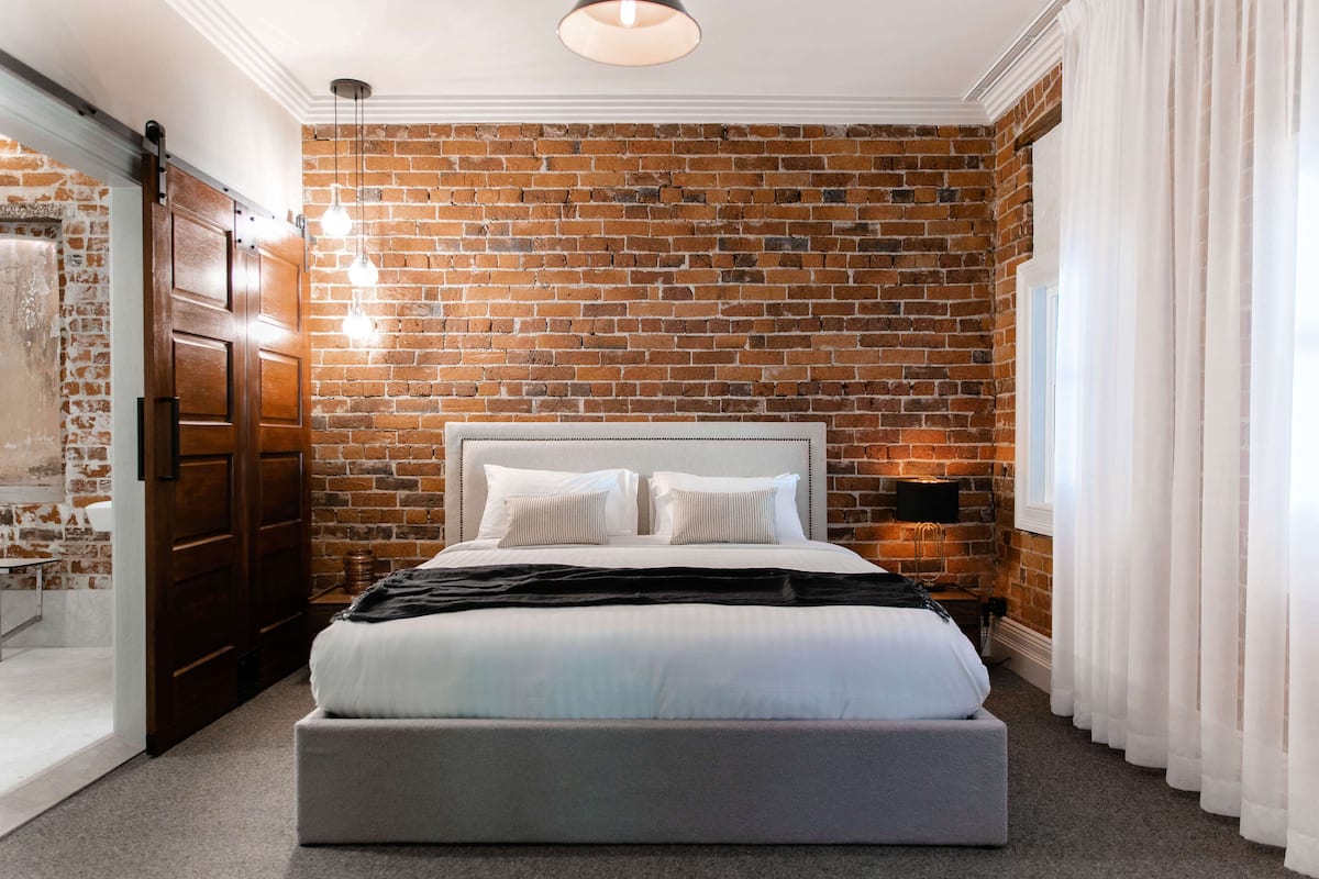 A bedroom with a brick wall, a large bed, a bedside table, and a window with curtains.