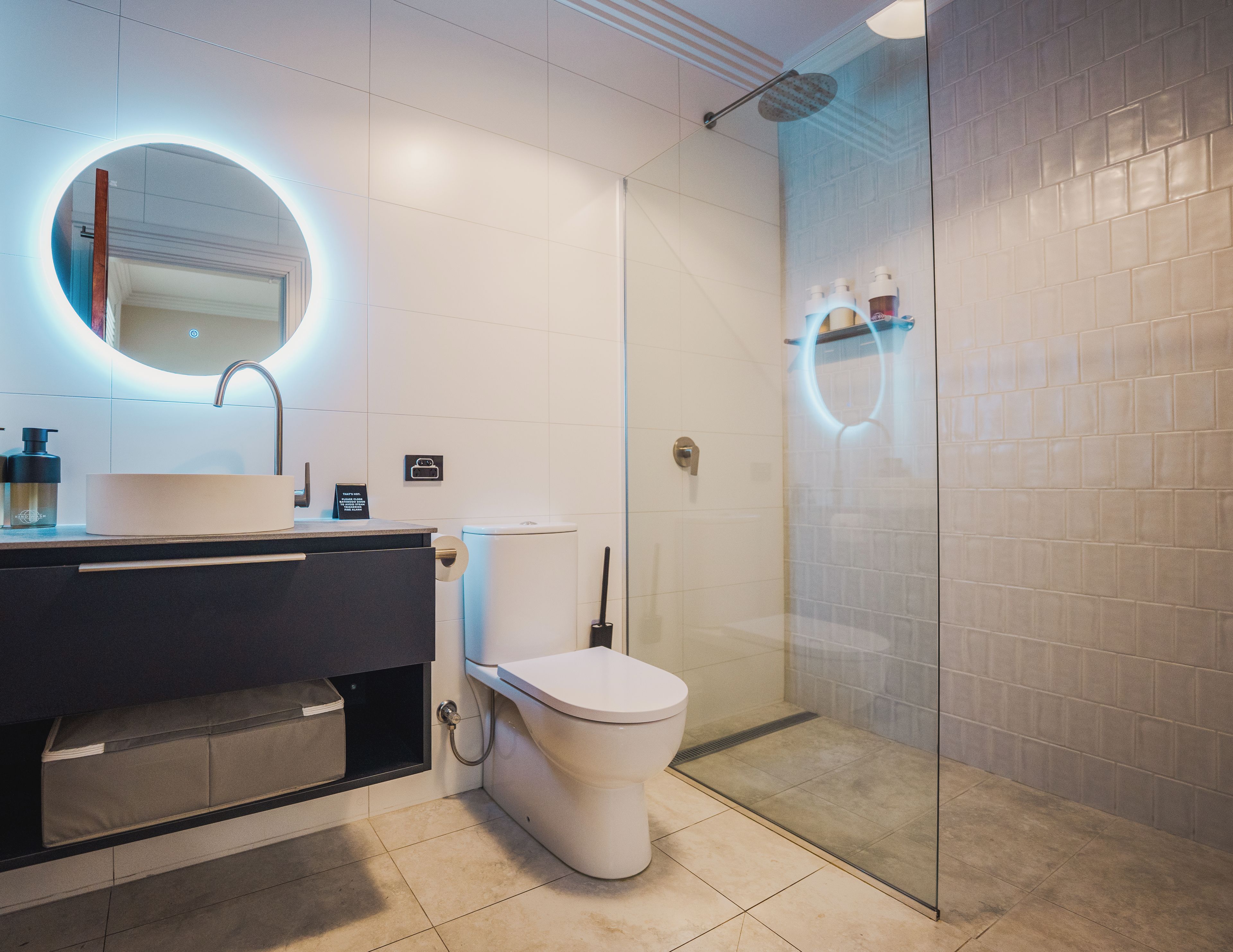 Un baño moderno con un espejo redondo, un inodoro y una zona de ducha con mampara de vidrio.