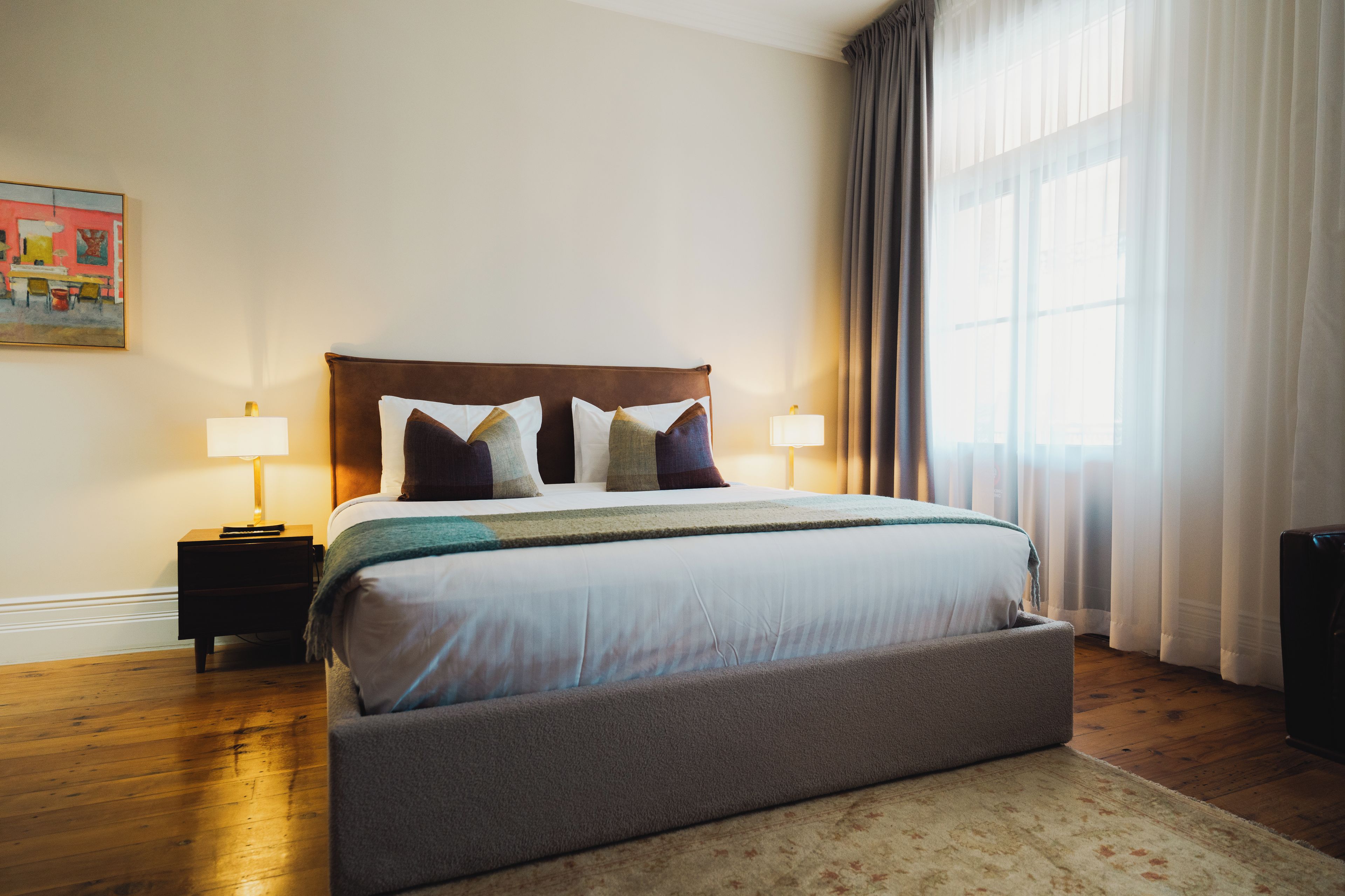 A neatly arranged hotel room with a large bed, two bedside tables, a nightstand, a window with curtains, and a painting on the wall.