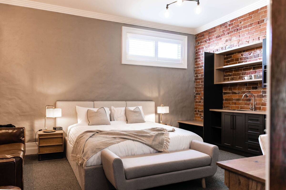 A bedroom with a large bed, bedside tables, a bench, a desk, and a brick wall.