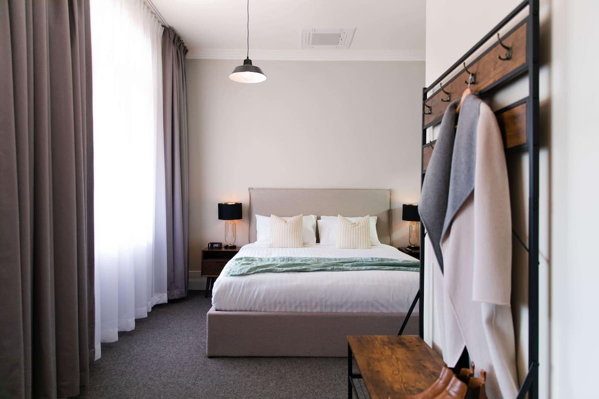 A hotel room with a large bed, a wooden bench, a coat rack, and a bedside table with a lamp.