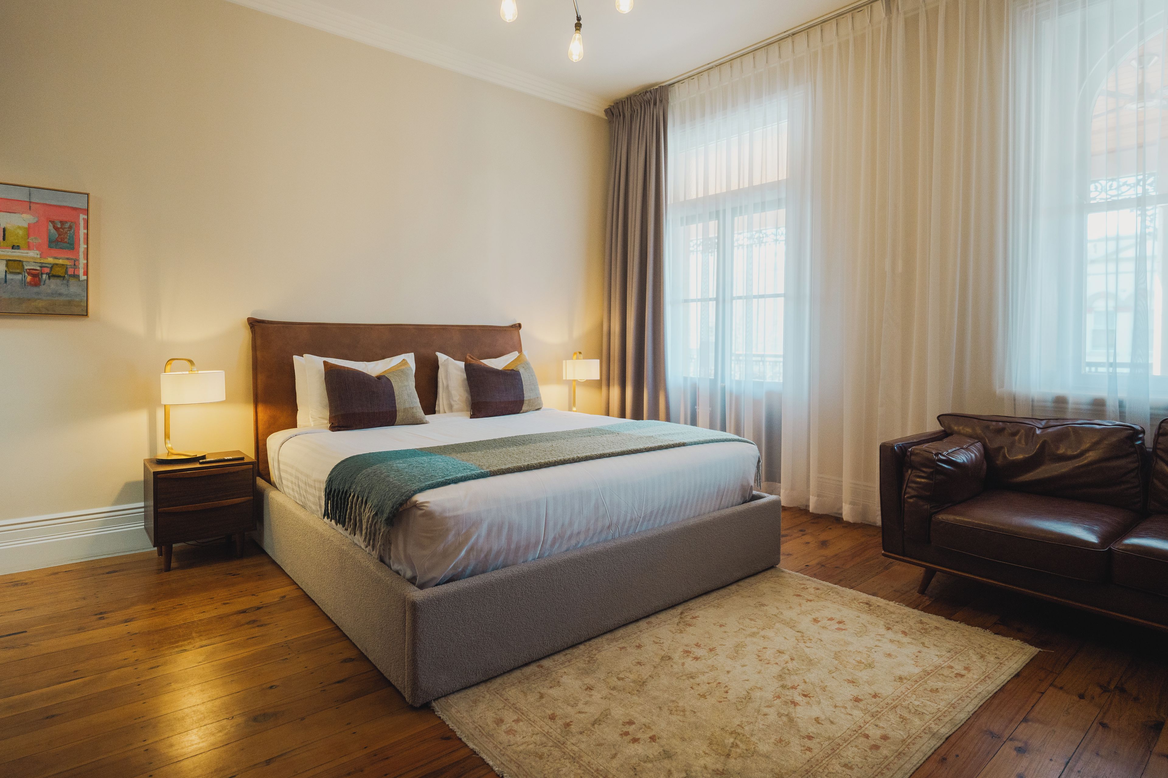Una habitación de hotel con una cama grande, una mesita de noche con lámpara, un sillón de cuero y una ventana con cortinas.