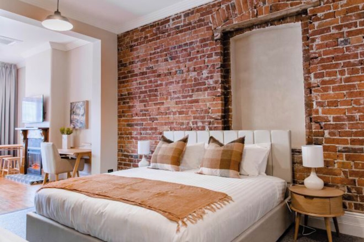 A hotel room with a brick wall, a bed with white bedding, a desk with a chair, and a lamp on a side table.