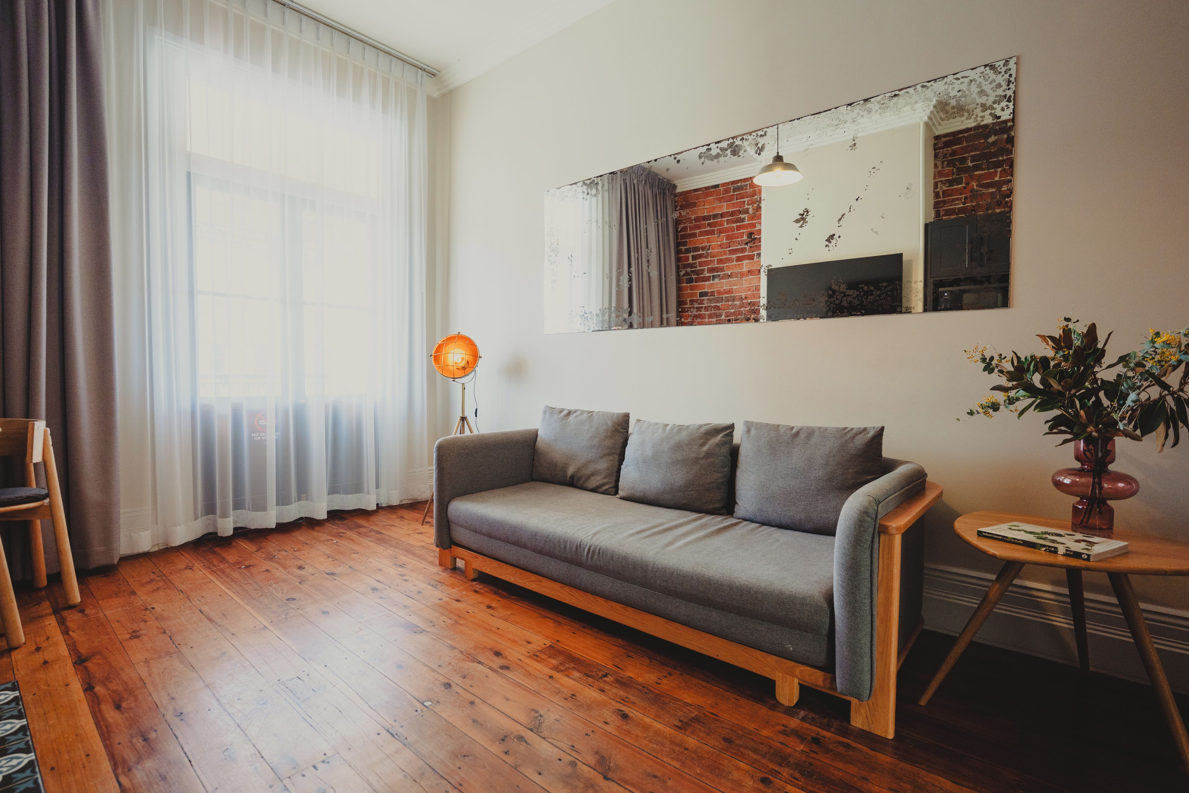 A living room with a grey sofa, wooden flooring, a lamp, a mirror, and a plant.