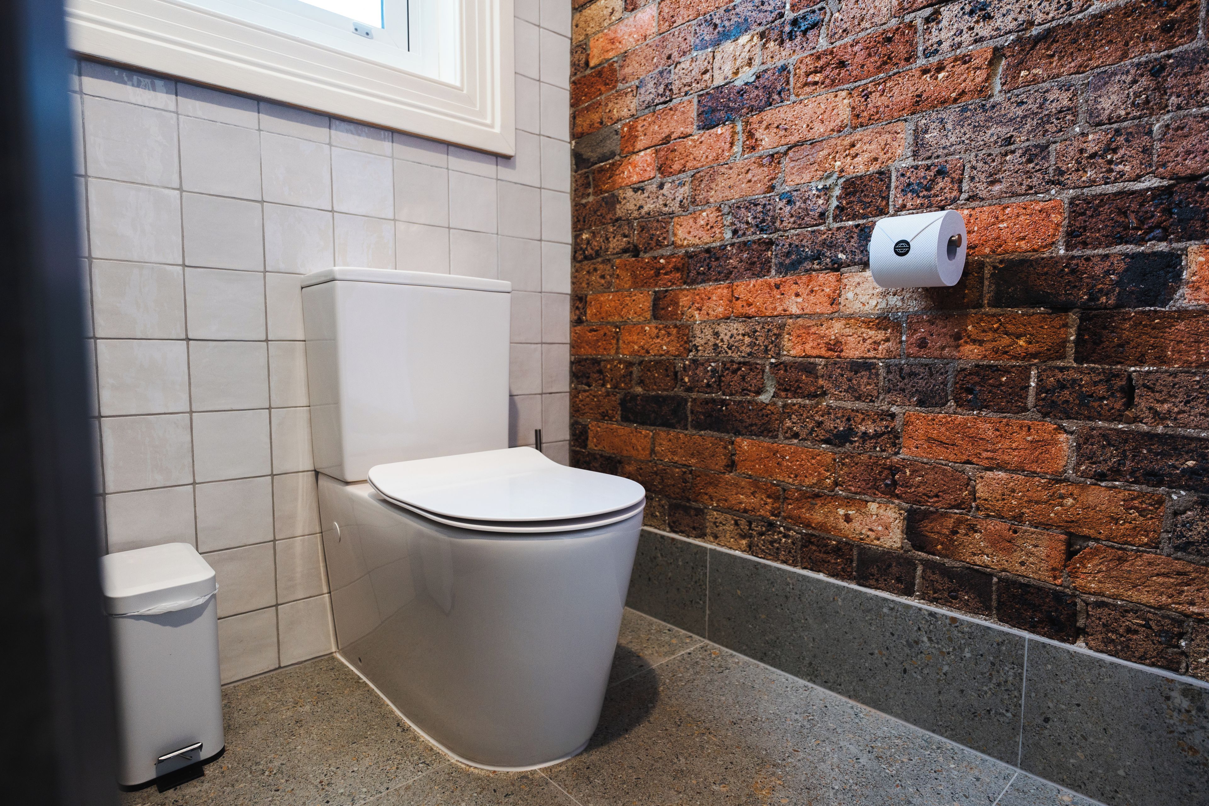 Une salle de bain moderne avec des toilettes blanches, une poubelle et un mur en briques.