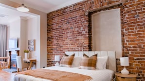 Un dormitorio con pared de ladrillo, una cama con ropa de cama blanca, una mesita de noche con lámpara y un escritorio con silla.