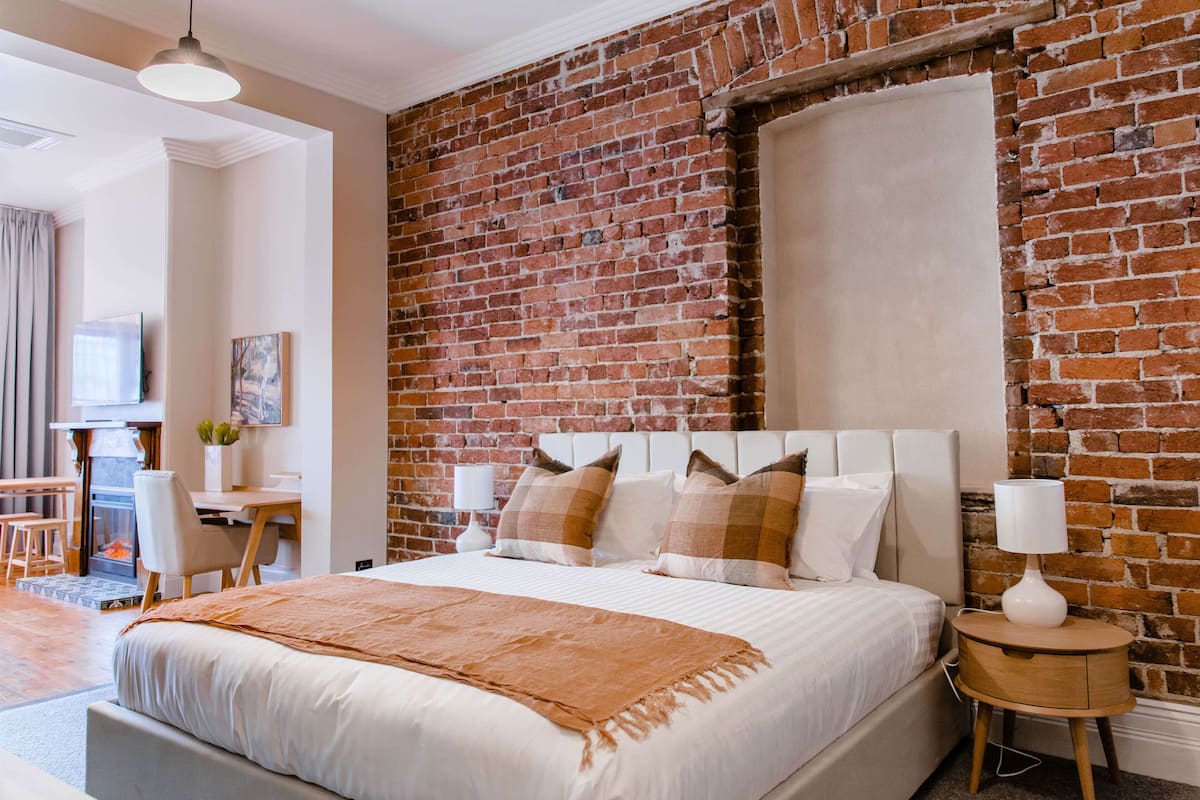 A bedroom with a brick wall, a bed with white bedding, a bedside table with a lamp, and a desk with a chair.