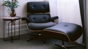 Una habitación de esquina con un sillón de cuero negro, una mesita auxiliar con una planta, una pared de ladrillo y una ventana con cortinas de gasa.