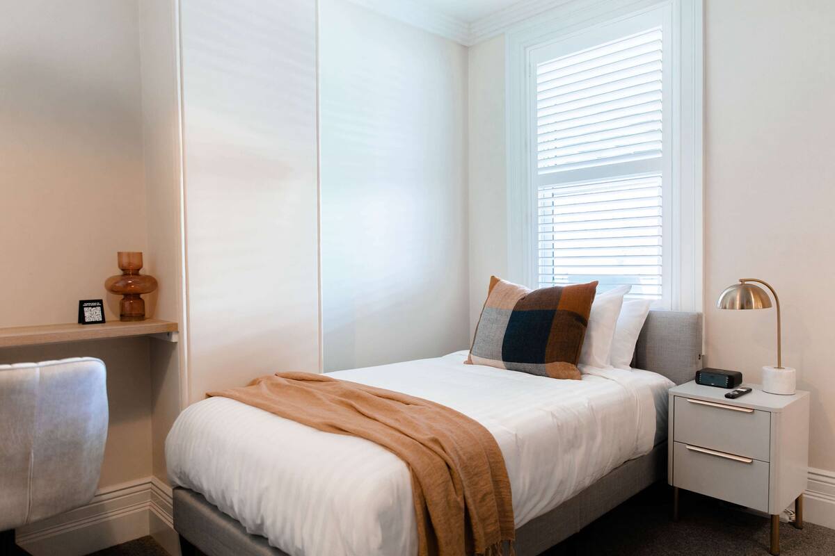 A neatly arranged bedroom with a bed, bedside table, lamp, and a window with blinds.