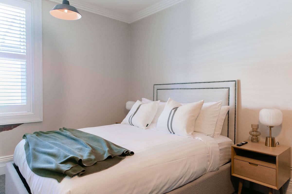 A neatly made bed with white linens and a green throw blanket, a bedside table with a lamp, and a window with blinds.