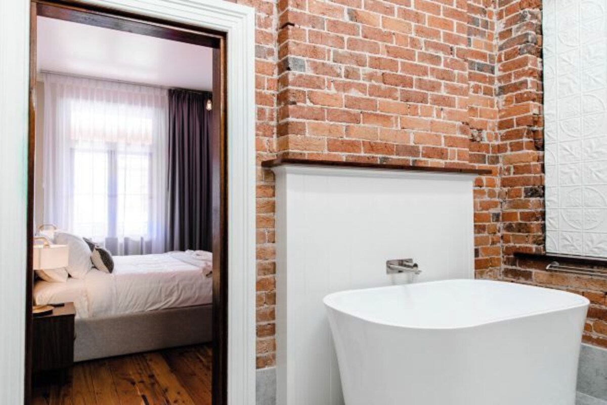 A modern hotel room with a brick wall, a large white bathtub, and a neatly made bed with a nightstand.