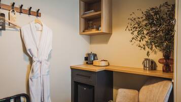 A hotel room with a robe hanging, a wooden shelf with books, a plant, a coffee maker, and a chair.