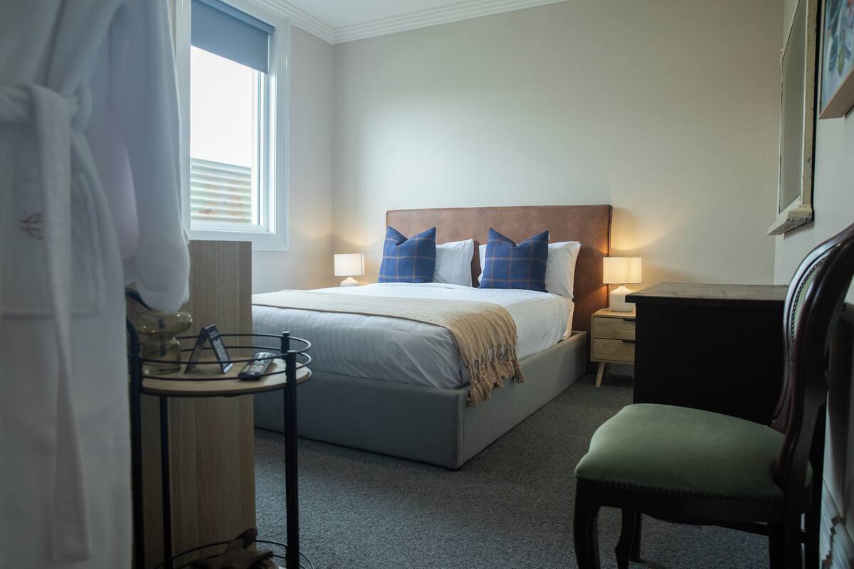 A hotel room with a bed, bedside lamps, a chair, a small table with a lamp, and a window with blinds.