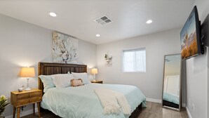 A bedroom with a bed, bedside tables, a wall-mounted TV, a window, and a ceiling vent.