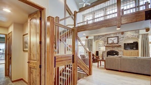 A spacious living area with a wooden staircase, a stone fireplace, and a large TV.