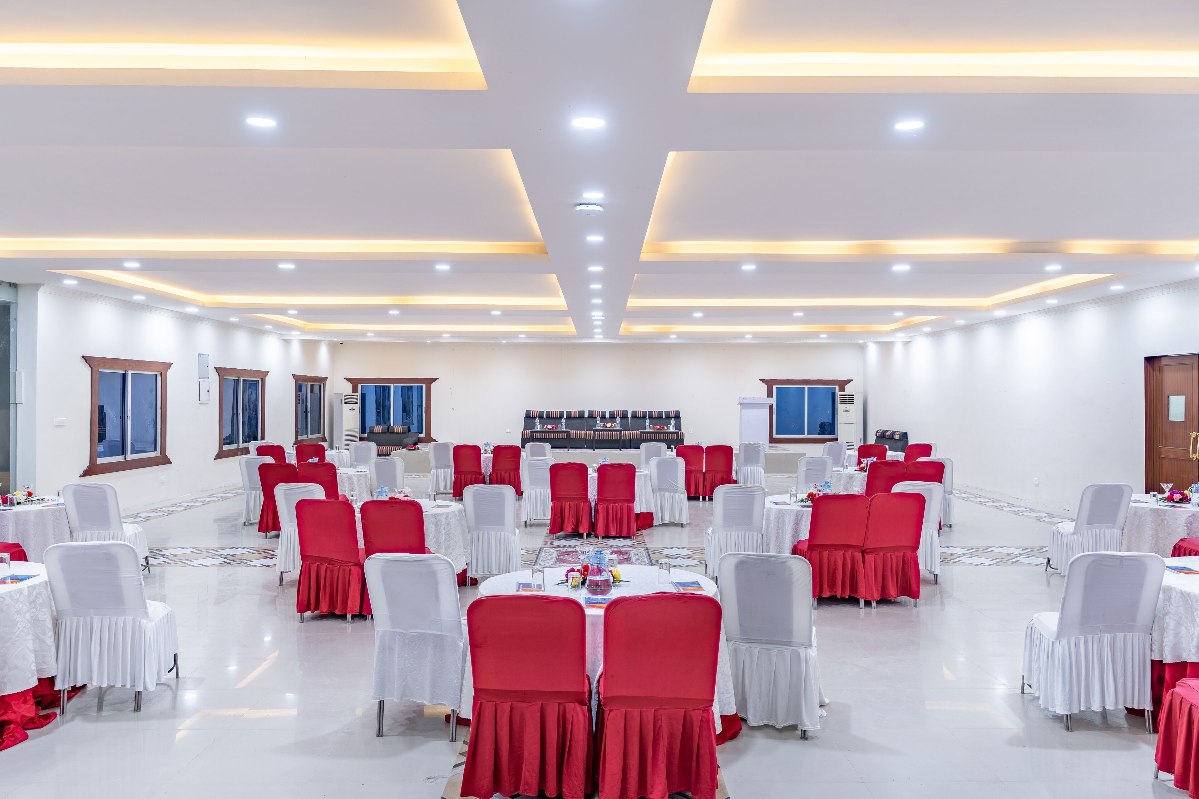 Un salón de banquetes con mesas redondas, sillas rojas y blancas, y un escenario con altavoces.