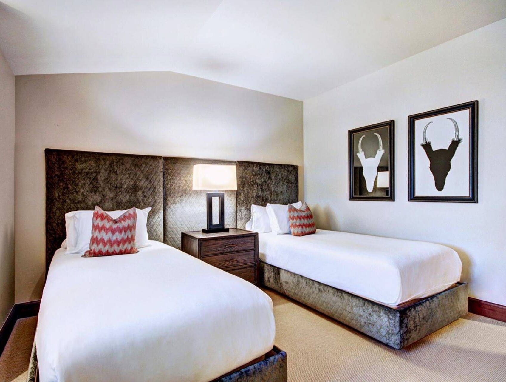 A hotel room with two beds, a nightstand with a lamp, and two wall-mounted pictures of animal horns.