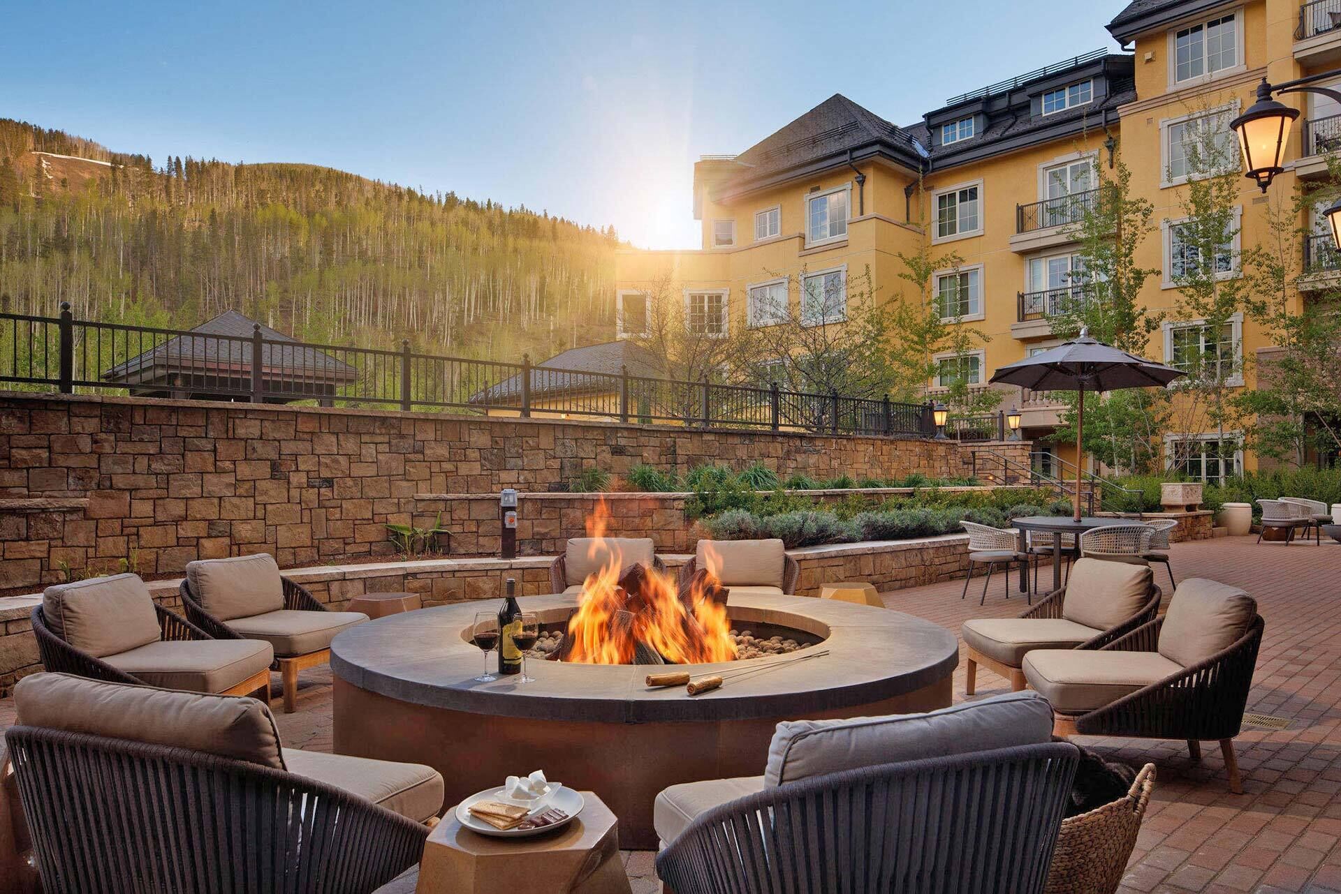 A fire pit surrounded by outdoor seating, a stone wall, and a building with balconies and windows in the background.