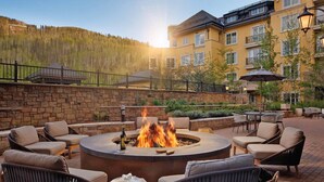 A fire pit surrounded by outdoor seating, a stone wall, and a building with balconies and windows in the background.