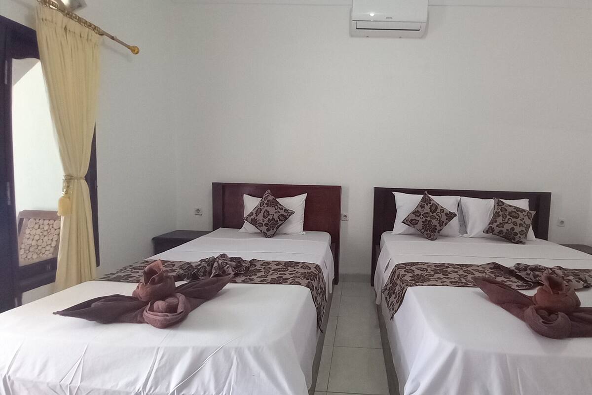 A hotel room with two single beds, each with a wooden headboard, white bedding, and decorative pillows. There is a small bedside table with a lamp, a wall-mounted air conditioner, and a curtain on the left side.