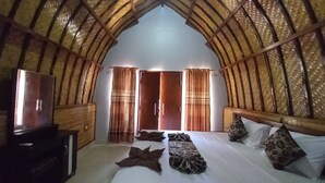 A room with a high, vaulted ceiling, a bed with pillows and a blanket, a television on a wooden stand, and a wardrobe. - Joevana Cottage (Gili Trawangan)