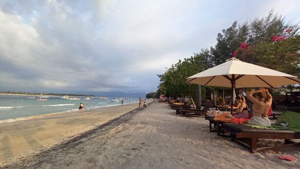 A beachside area with a sandy walkway, umbrellas, and wooden benches. - Joevana Cottage (Gili Trawangan)