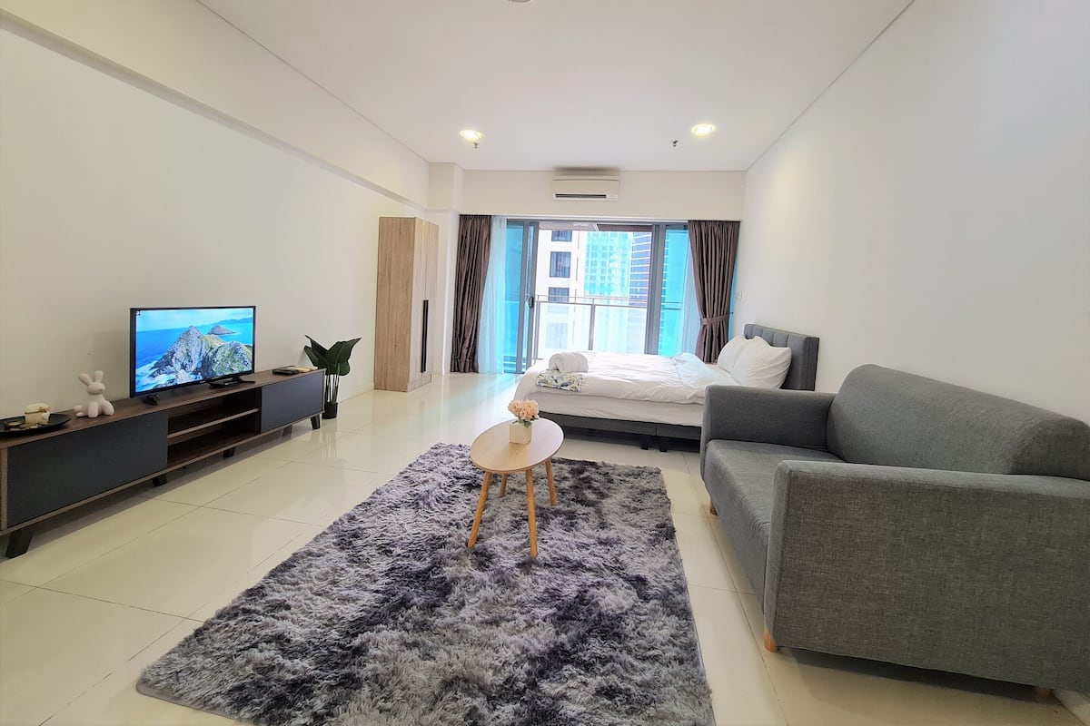 A modern hotel room with a bed, a grey sofa, a small table, a TV, and a large window with curtains.